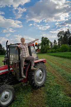 A smiling blonde woman in a plaid shirt poses joyfully while standing on the steps of a red tractor in a green field, woman farmer concept
