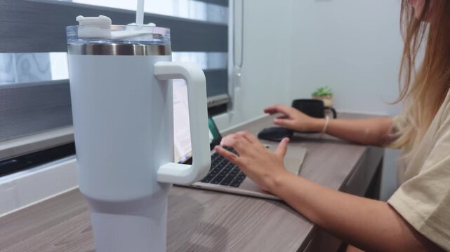 Woman engaging in remote work, sitting at a desk and typing on a laptop, with a large reusable tumbler emphasizing daily hydration and a focused workspace environment
