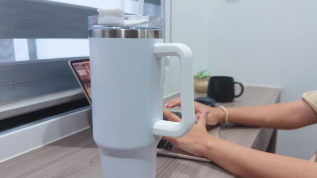 Insulated tumbler on a desk with a person working on a laptop, emphasizing hydration and productivity in a modern remote work or office environment