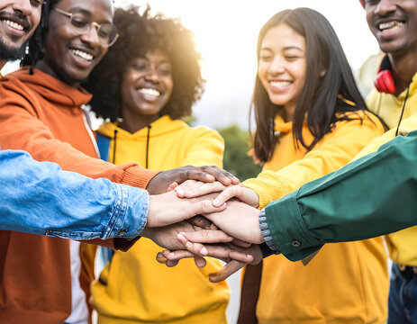 Diverse group of young friends stacking hands in a huddle, showing unity and teamwork, outdoors with warm sunlight