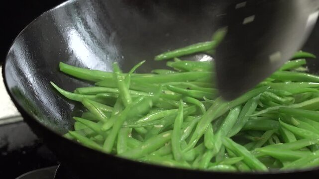 Fresh green beans stir-fried in black carbon steel wok with metal spatula, close-up cooking action