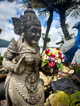 Ornate carved stone Balinese deity statue with floral offerings and a peacock sculpture at Ulun Danu Beratan Temple, Lake Bratan, Bedugul, Bali, Indonesia