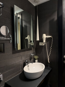 Stylish compact hotel bathroom with dark wall tiles, white ceramic vessel basin, rectangular mirror, wall-mounted hairdryer, chrome fixtures and a small decorative plant