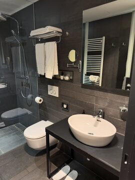 Contemporary hotel bathroom featuring dark charcoal wall tiles, a white vessel sink, glass shower enclosure, wall-mounted toilet, large mirror, heated towel rail and neatly folded white towels