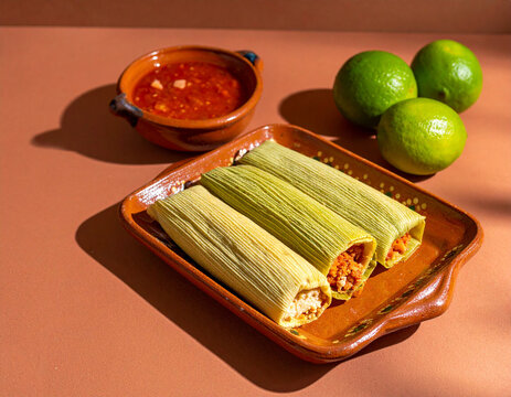 Steaming tamales wrapped in corn husks sit on a terracotta tray next to a bowl of spicy red salsa and limes.