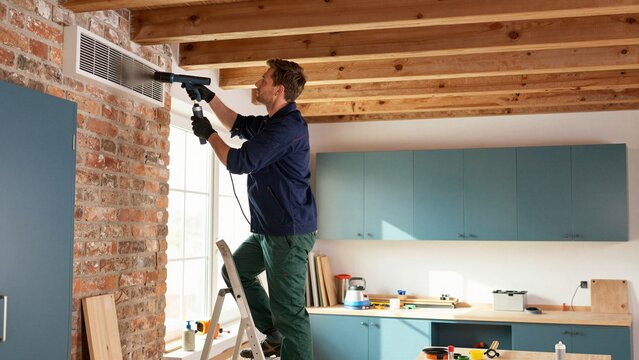 A technician performs ventilation maintenance by cleaning an air duct with a powerful nozzle in a modern kitchen setting.