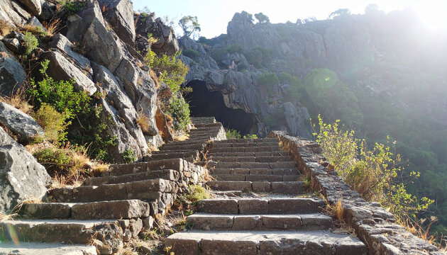 Old carved stone steps integrated into rocky mountain slope, leading toward shadowy natural cave, aged architecture blending seamlessly with rugged stone and vegetation.