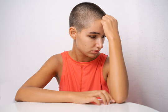 Young caucasian woman with a shaved head in an orange tank top touching her forehead at a white table, for headache, migraine and fatigue content