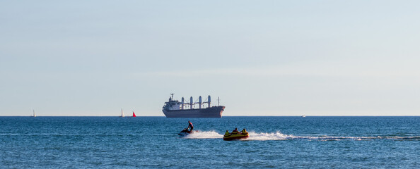  jet ski towing yellow inflatable tube under clear sky. large cargo ship with deck cranes with a damaged wheelhouse as a result of a drone attack, sailboats visible on horizon © Andrey Solovev