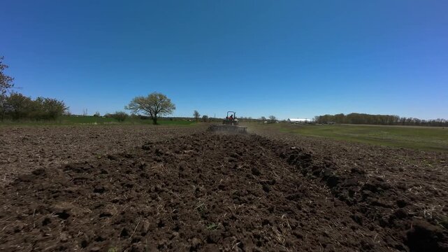 Low angle aerial view tracking forward toward a green utility tractor pulling a disc harrow attachment across deep textured dirt soil in Milton Ontario under a clear blue sky.