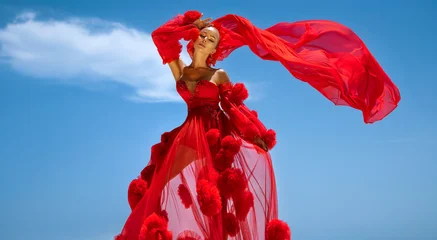 Glamorous fashion model in a dramatic red tulle dress posing on white architecture against a luxury tropical resort backdrop with palm trees and blue sky © marcink3333