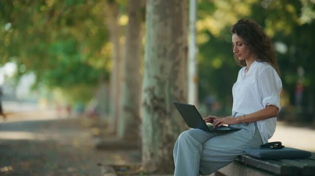A focused businesswoman works on her laptop in a serene park environment. She conducts an outdoor business meeting, connecting with her tasks amidst nature.