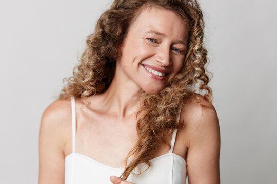 An authentic middle-aged woman with natural skin and no retouching smiles happily, holding her curly hair. Concept for real beauty, aging gracefully, and self-acceptance.
