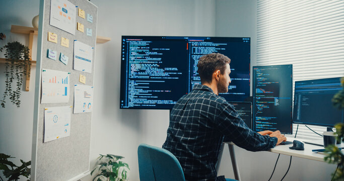 Young Caucasian male programmer writes code using multiple screens in home office workspace. AI development, cybersecurity, network database system, software engineering, remote tech startup concept.