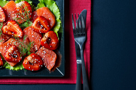 Gourmet salmon and strawberry salad appetizer on black plate, closeup. Top view of elegant salmon carpaccio with fresh strawberries sesame seeds and dill served on dark background