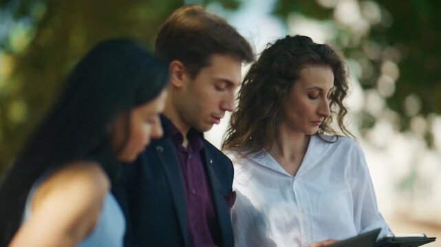 Three young colleagues engage in an outdoor discussion, reviewing documents and a tablet. They collaborate on a project, focused and concentrated in a green environment.