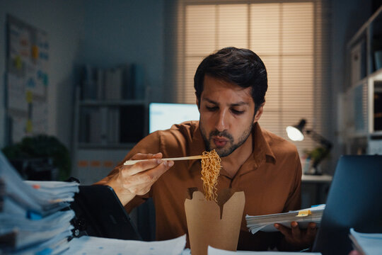 Young Latin businessman eating takeout instant noodles and reviewing document on laptop at desk in dark night office. Overtime working late, overworked deadline, fast food, corporate problem concept.