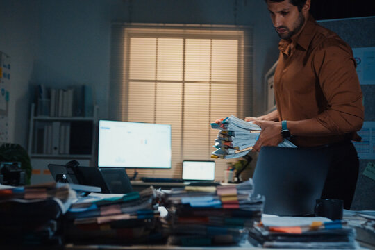 Stressed young Latin businessman walking to place tax document stack at desk in dark night office. Overworked overtime, heavy workload stress, tired employee, corporate business burnout concept.