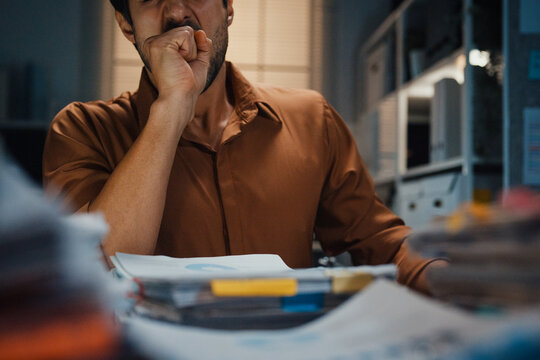 Sleepy young Latin businessman yawning over heavy paperwork stack at desk in dark night office. Overwork burnout, working late overtime, corporate job stress, exhausted professional manager concept.