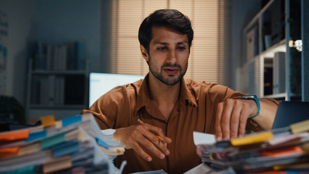 Overworked young Latin businessman working late on document in large stack of paperwork files at office night. Tax audit deadline, accounting stress, corporate burnout, urgent business concept.