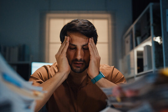 Stressed young Latin businessman having severe headache and migraine at desk in night office. Debt crisis, corporate burnout, late overtime workload, mental health anxiety, business failure concept.