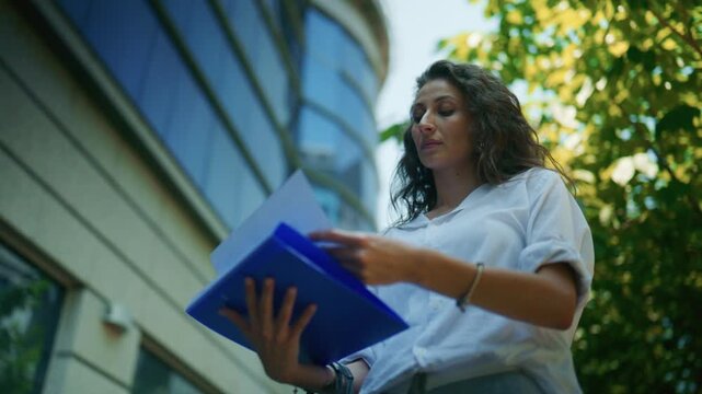 A young business-minded woman thoughtfully reviews documents in a blue folder outside an office building. She appears focused on her tasks in the urban environment.