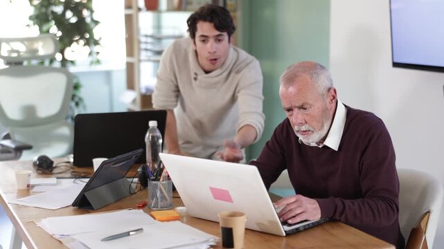 Senior man leaned over to seated guy employee, supervise activities, helps with work and looks at laptop screen. Travel agency, engineering company, notary office or legal advice