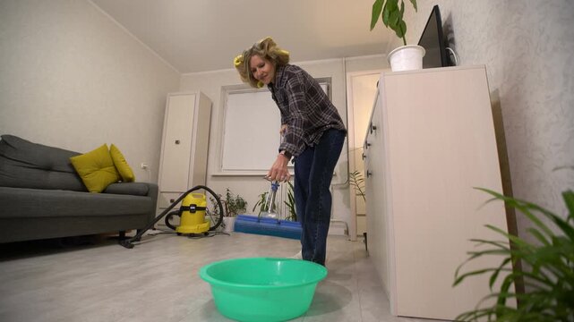 An elderly woman wearing a plaid shirt and jeans is seen wringing out a mop into a green basin on the floor. The living room features a grey couch with yellow pillows, a vacuum cleaner, and potted pla