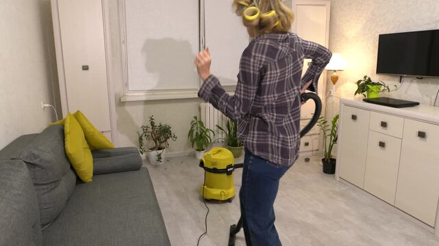 An elderly woman with curlers vacuums and dances into the living room. She is wearing a plaid shirt and jeans, and in the background there are plants, a gray sofa with yellow pillows and a lamp.