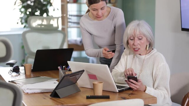 Adult woman sternly scolds elderly female colleague for using mobile phone while working in office