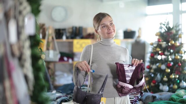 Woman client looks at products in store, looks closely at bra, picks up wardrobe item. Concept of seasonal wardrobe shopping. Young woman enjoys shopping for stylish clothes 