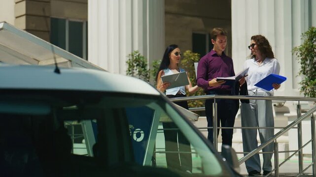 Three business people hold documents and discuss work matters outside a modern building. A small team collaborates on a project outdoors on a sunny day.