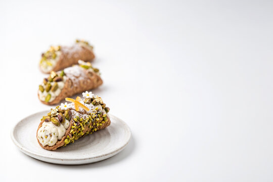 Traditional Sicilian cannolo garnished with abundant chopped pistachios, candied orange peel, tiny flowers, and gold leaf. Exquisite classic Italian dessert presented on a ceramic saucer.