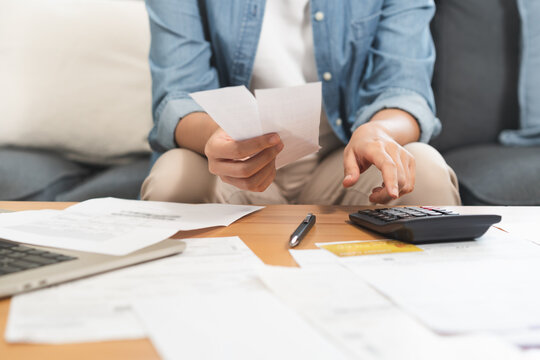 Stressed asian young business woman, employee using calculator to calculate expenses of monthly, hand holding bills and receipt for to payment on table at home. Financial, finance of banking concept.