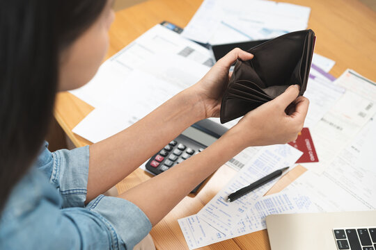 Stressed person opening empty wallet with no money inside surrounded by unpaid bills and expenses, suffering from financial difficulties, loan debt and household budget problems indoors.