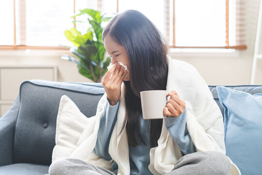 Sick concept, tired asian young woman have a fever, flu and hand in use tissues paper sneezing nose, runny and holding hot mug  while sitting on sofa at home. Health care with disease, pandemic virus.
