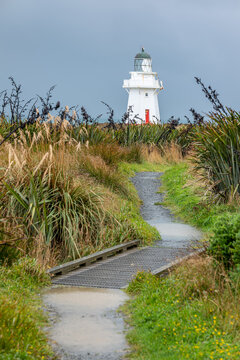 Waipapa Point Lighthouse mit roter T&uuml;r auf Grasd&uuml;nen vor st&uuml;rmischer See und dunklem Wolkenhimmel, Southland, Neuseeland