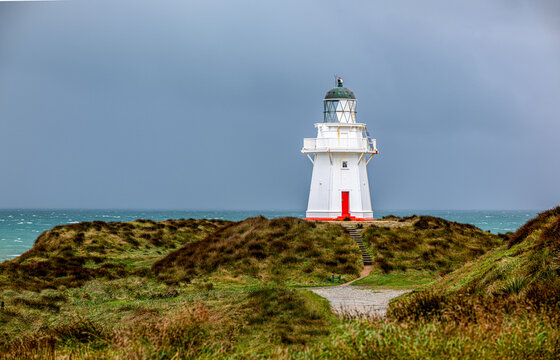 Waipapa Point Lighthouse mit roter T&uuml;r auf Grasd&uuml;nen vor st&uuml;rmischer See und dunklem Wolkenhimmel, Southland, Neuseeland