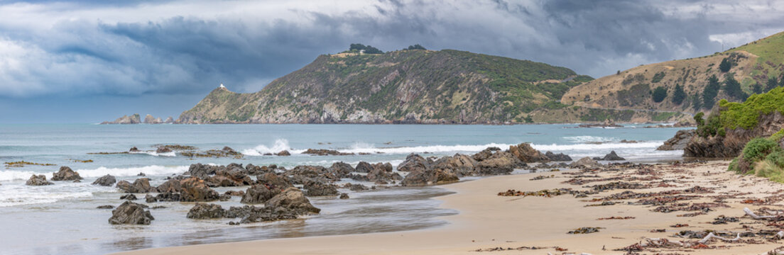 Sandstrand am Kaka Point Beach mit bewaldeter Klippe, Treibholz und Brandung unter Wolkenhimmel, Otago, Neuseeland
