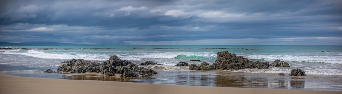 Sandstrand am Kaka Point Beach mit bewaldeter Klippe, Treibholz und Brandung unter Wolkenhimmel, Otago, Neuseeland