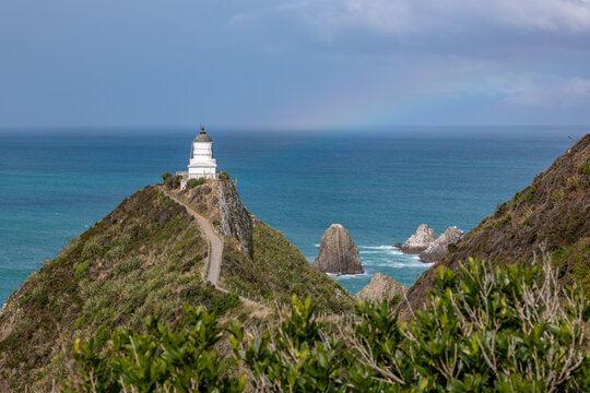 Nugget Point Lighthouse auf schmalem Felsgrat mit Blick auf die Nuggets-Felsen und schwachem Regenbogen, Otago, Neuseeland
