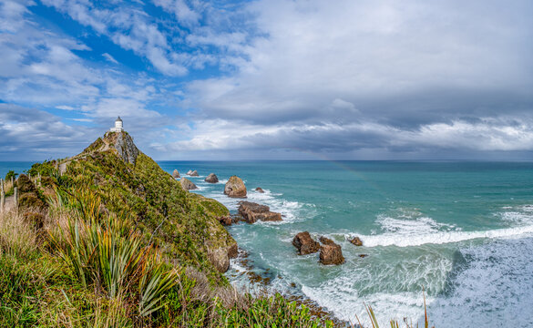 Nugget Point Lighthouse auf schmalem Felsgrat mit Blick auf die Nuggets-Felsen und schwachem Regenbogen, Otago, Neuseeland