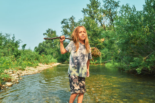 Boy standing in a river with a stick and clump of weeds over his shoulder