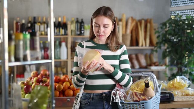 Smiling young girl with shopping basket trying to make choice holding two sorts of cheese in grocery store with varied stock