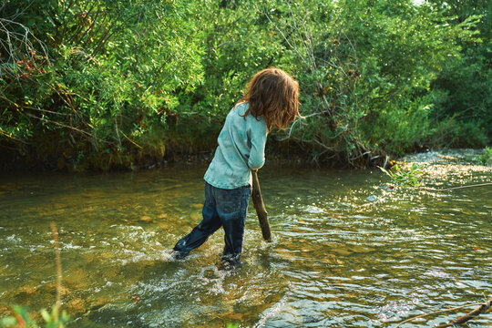 Boy wading through a river holding a wooden stick