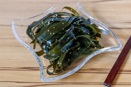 Seasoned seaweed kelp salad sprinkled with white sesame seeds served in transparent glass dish on wooden table with chopsticks, healthy Asian appetizer concept
