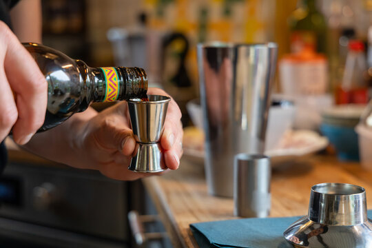 A person pours a dark liqueur into a metal jigger while preparing cocktails in a kitchen.