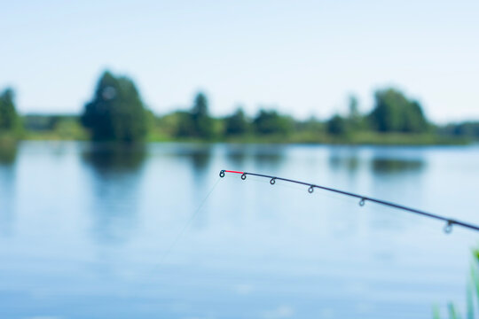 Fishing feeder rod tip over calm lake water with blurred natural landscape background. Outdoor angling scene for fishing brands, recreation marketing, sports equipment advertising and lifestyle.
