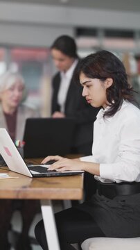 Asian girl sitting at desk is working on laptop computer among staff colleagues. Working day in office of business structure, manager is working at computer. 