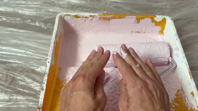 Woman's hands covered in light pink paint over a yellow paint tray during renovation. DIY home renovation.